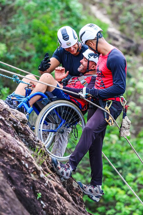 Free Three men, including one in a wheelchair, rappelling down a cliffside, showcasing inclusive adventure sports. Stock Photo