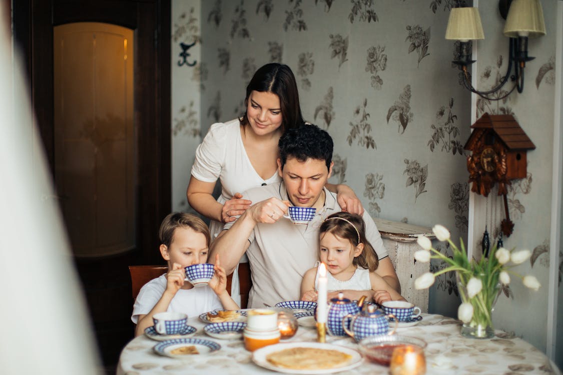Free Contented family drinking tea with crepes together while sitting at table with candles at cozy kitchen with cuckoo clock on wall Stock Photo