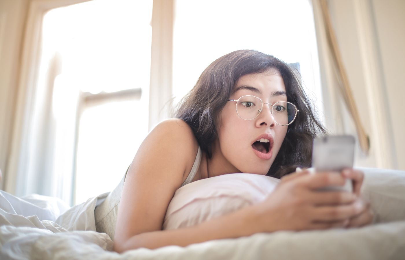 Young woman lying on bed, surprised by something on her smartphone, in a bright bedroom setting.