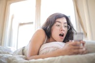 明亮的卧室里，一位年轻女子躺在床上，惊讶地看着手机上的内容