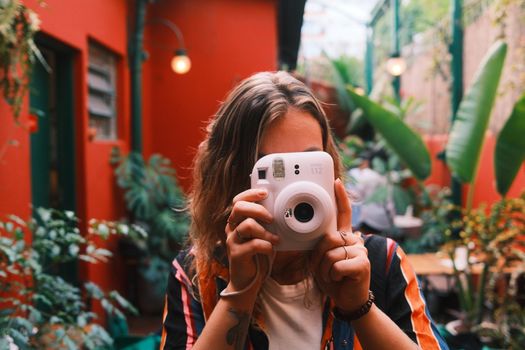 A young woman holds a camera in a colorful urban café with lush plants, capturing a lively moment.