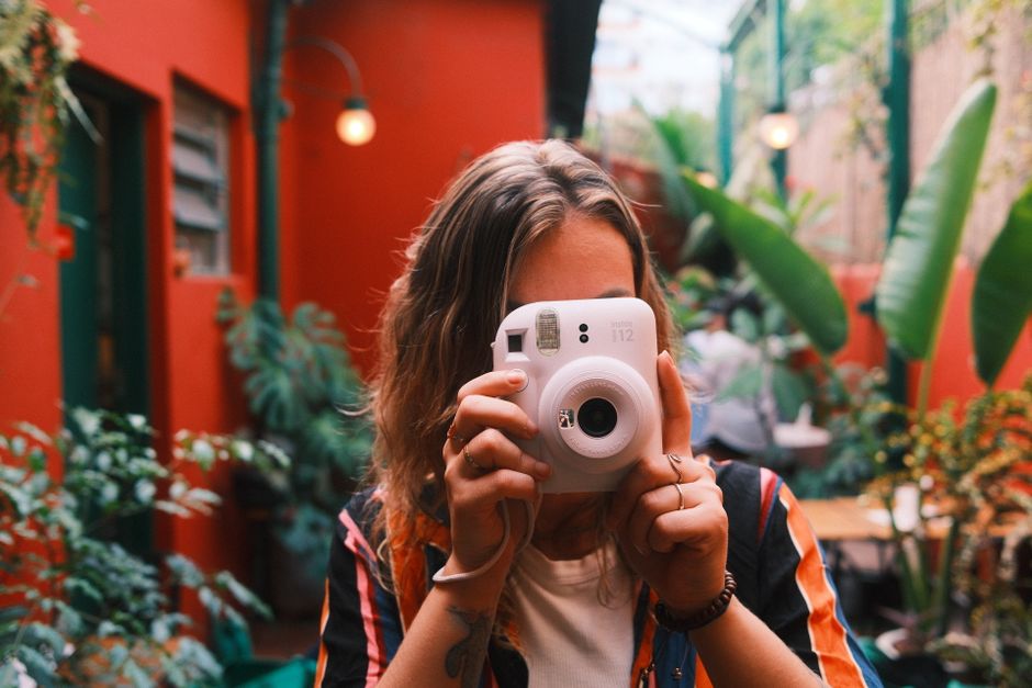 A young woman holds a camera in a colorful urban café with lush plants, capturing a lively moment.