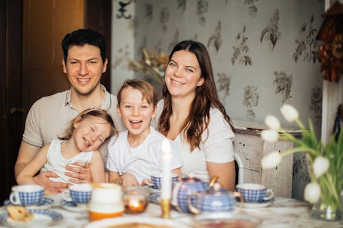 Positive family in casual clothes enjoying tea with sweets sitting at table set for breakfast with candle and laughing while looking at camera