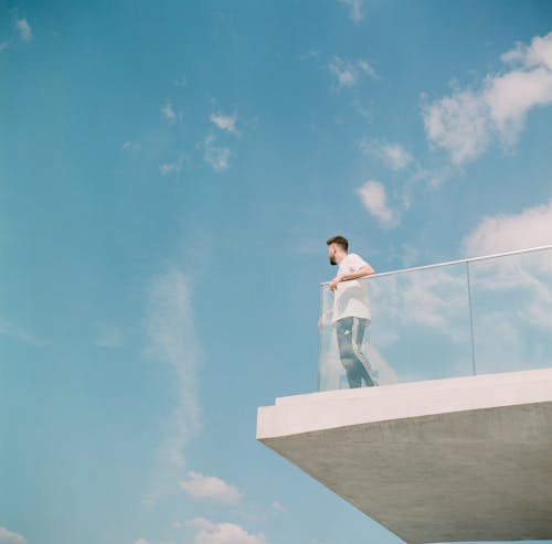 Man in White Long Sleeve Shirt and Joggers Standing on Balcony