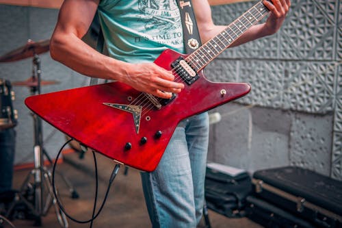 Crop male musician in tee shirt and jeans standing and playing red electric guitar with drums behind