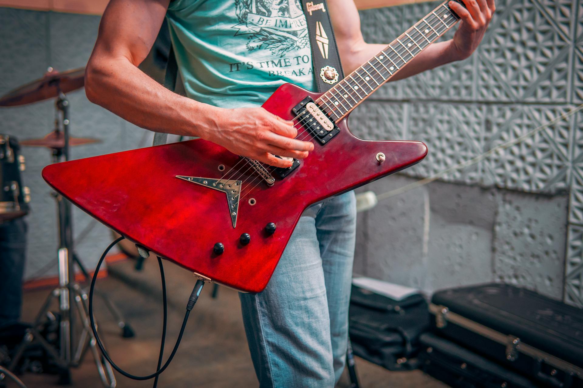 Crop male musician in tee shirt and jeans standing and playing red electric guitar with drums behind