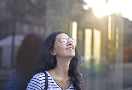 Free A woman with eyeglasses and striped shirt smiling serenely while leaning against a glass wall outdoors. Stock Photo