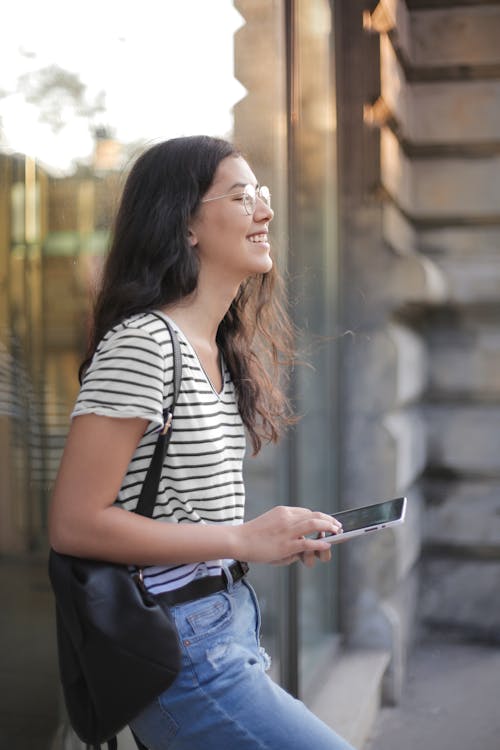 Side view of teen female in casual outfit and glasses browsing smartphone while smiling standing on street