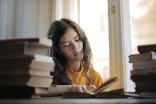 Young smart Asian woman reading book while sitting at table by window in soft daylight