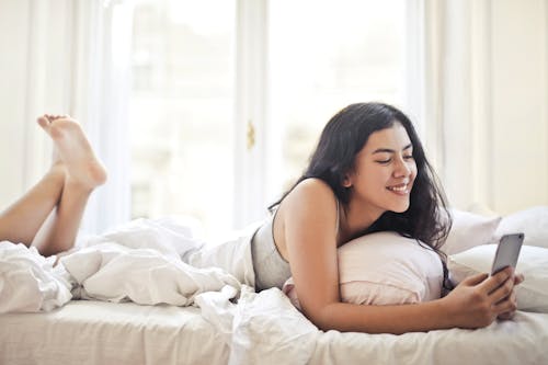 Free Happy young woman browsing phone on bed Stock Photo