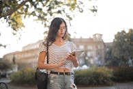 阳光明媚的公园里，一名穿着条纹衬衫和破洞牛仔裤的年轻女子正使用着平板电脑