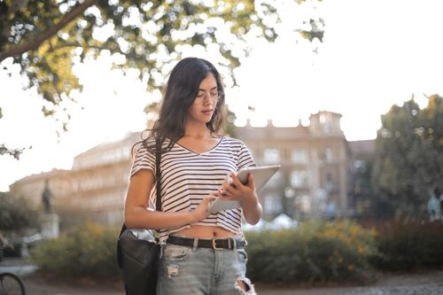 Free A young woman in a striped shirt and ripped jeans using a tablet in a sunny park. Stock Photo