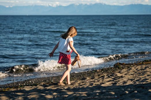 A young girl walks barefoot on a sandy beach in Victoria, Canada, enjoying a sunny day by the ocean.