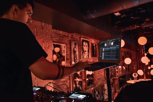 Male DJ in black tee shirt standing behind equipment and selecting songs on laptop during party in bar