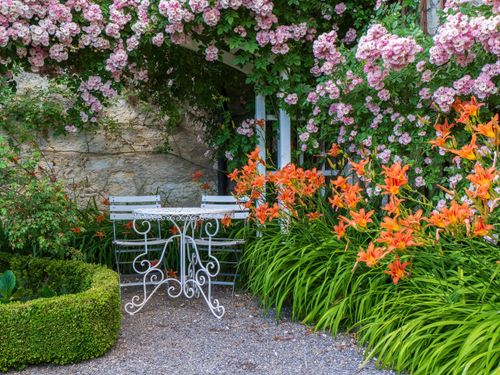 Free Enchanting garden setting with vibrant flowers and a classic wrought-iron table and chairs. Stock Photo