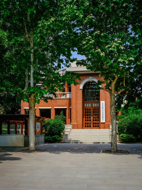 Free Front view of a historic red brick building surrounded by lush green trees on a sunny day. Stock Photo
