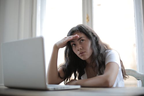 Free Irritated ethnic female entrepreneur in casual wear sitting at table with netbook and touching head while waiting for internet connection during remote work Stock Photo