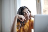 一位穿着黄色衬衫的女子在室内窗边若有所思地拿着一支笔