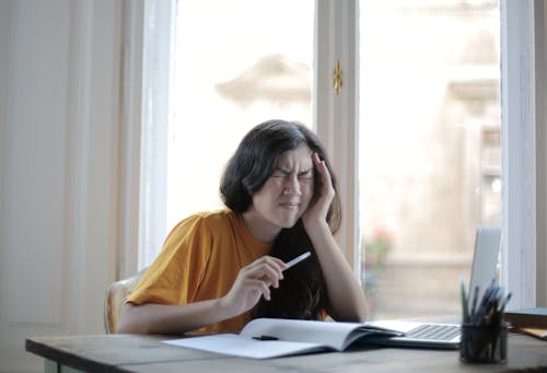 Free Ethnic female student in casual wear sitting at table with laptop and notebook and grimacing from migraine while doing homework during distance learning Stock Photo
