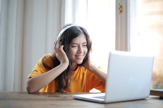 Woman in a yellow shirt enjoys music while working on a laptop at home.
