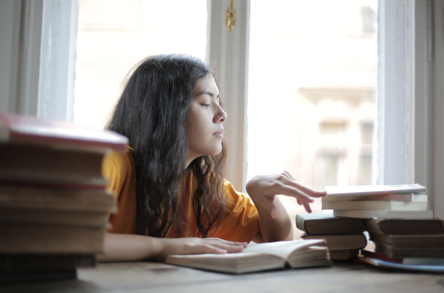 Side view of young ethnic woman in casual clothes sitting at table in living room and reading books while doing research and working on assignment
