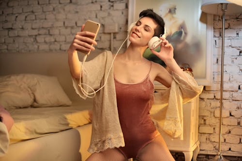 Gorgeous female in provocative wear and fishnet tights sitting on floor with closed eyes and enjoying songs while taking selfie on smartphone