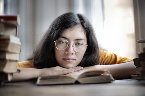 Exhausted ethnic female student in eyeglasses lying on table with stacks of textbooks while working on assignment and looking at camera