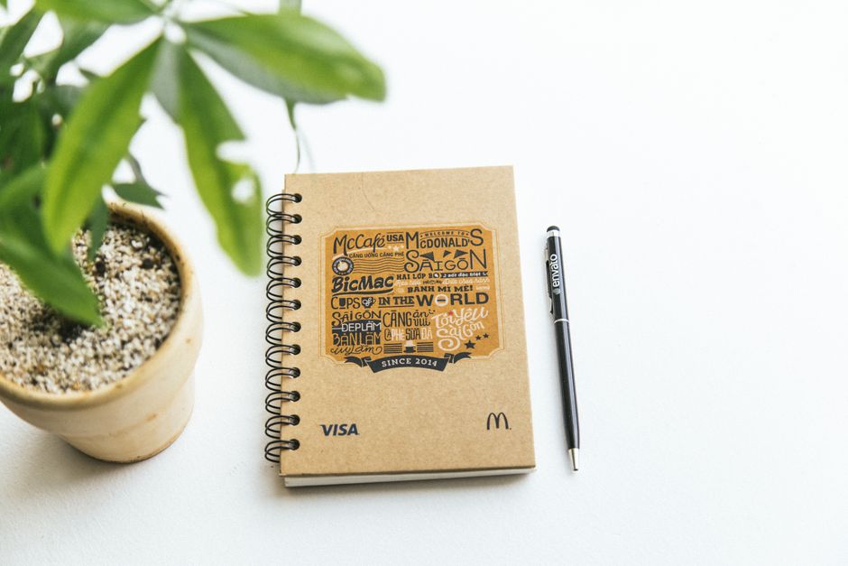 A spiral notebook and pen on a white background next to a potted plant, creating a minimalistic workspace vibe.