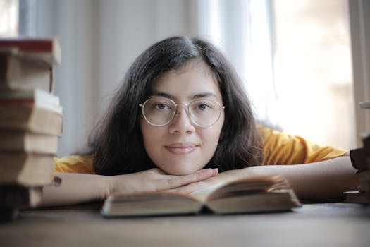 一位戴眼镜的女子在室内将下巴搁在一本书上，周围堆满了书