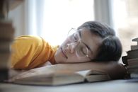 一名戴着眼镜的年轻女子趴在桌上睡着了，桌上摊开着一本书，看起来是学习累了在休息