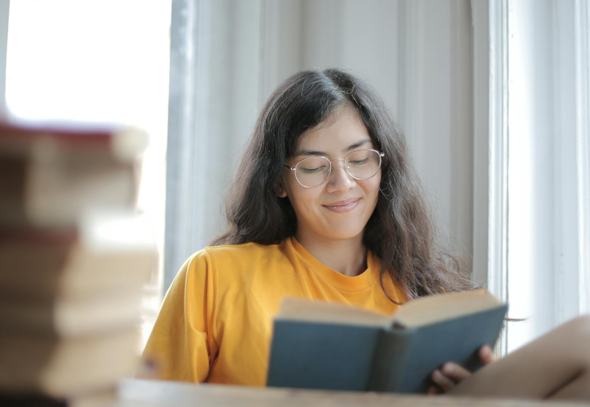 A smiling woman wearing a yellow shirt reads a book by a window indoors.. smiling, woman, wearing