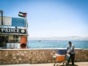 A man with a cart near a boat docked at the Red Sea in Aqaba, with scenic mountains in the background.