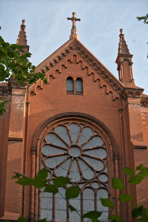 Free Close-up of a Gothic Revival church façade featuring a large rose window and spires, framed with greenery. Stock Photo