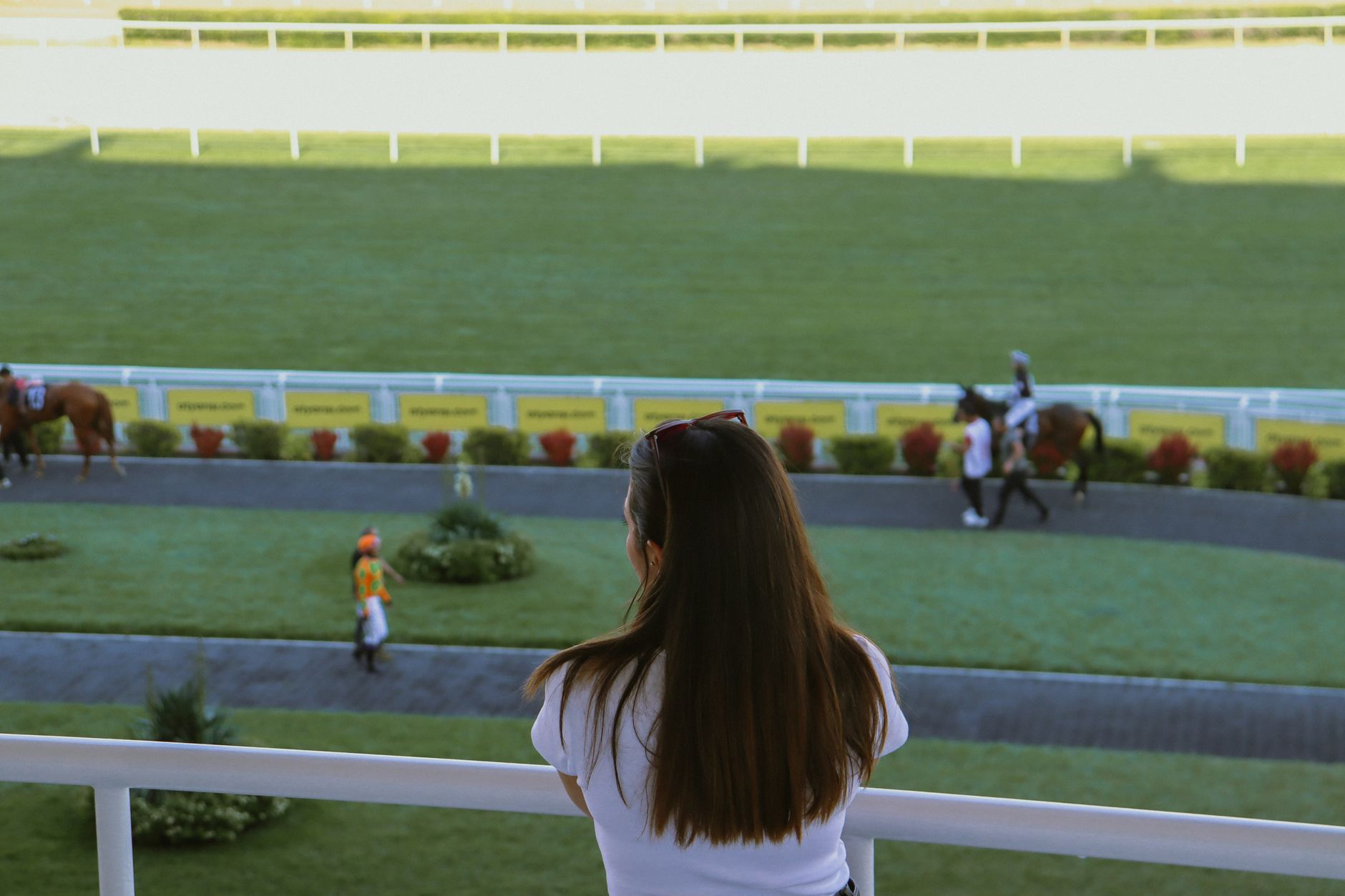 A woman gazes at a horse racetrack, observing jockeys and horses preparing for a race.