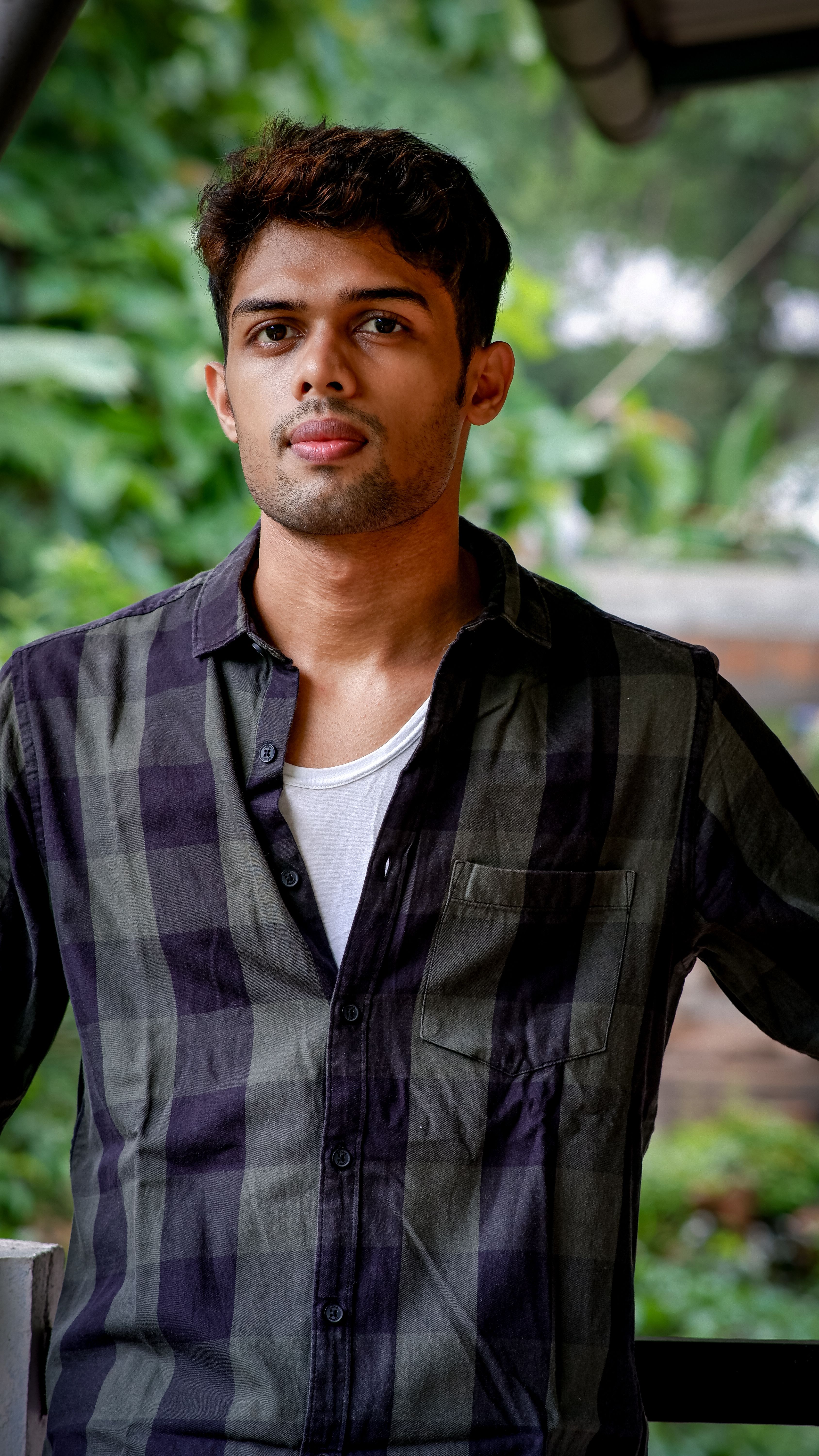 A young man in casual attire posing outdoors with a lush green background.