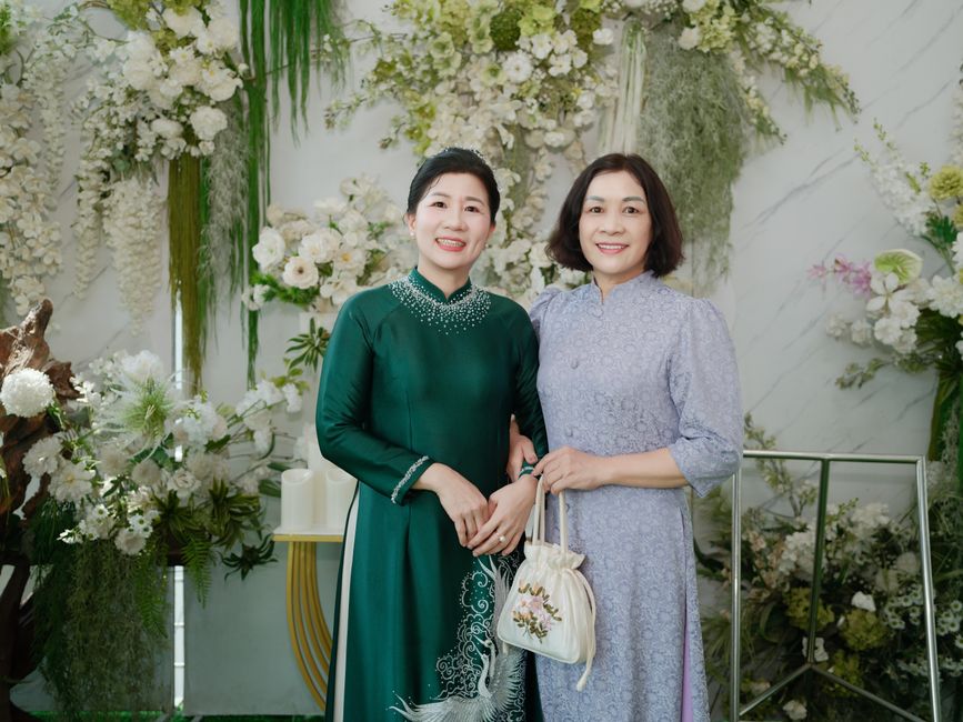 Two women in traditional dresses pose in front of a floral backdrop, conveying elegance and cultural richness.