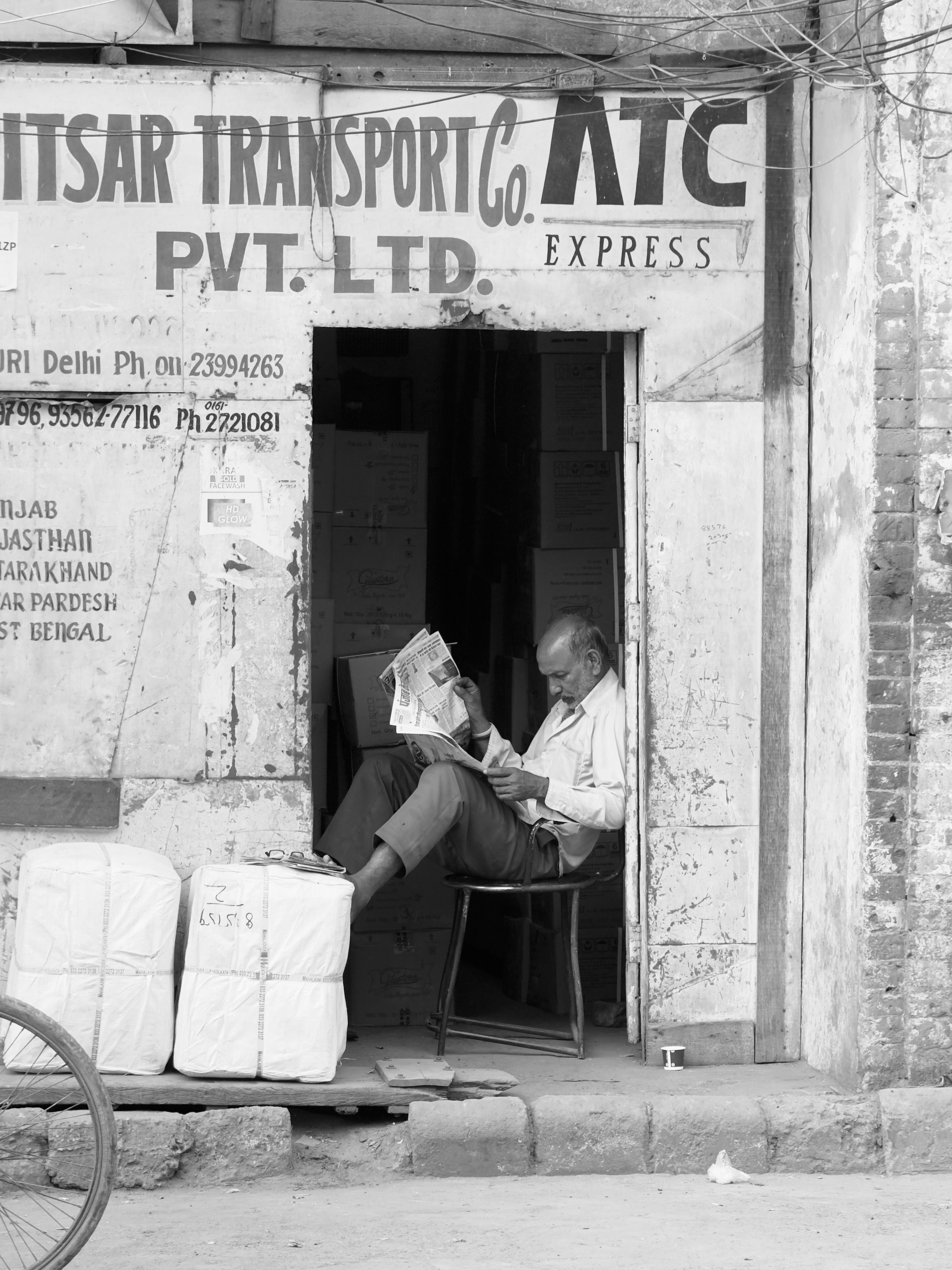 Black and white photo of a man reading newspaper in a street-side doorway in India.