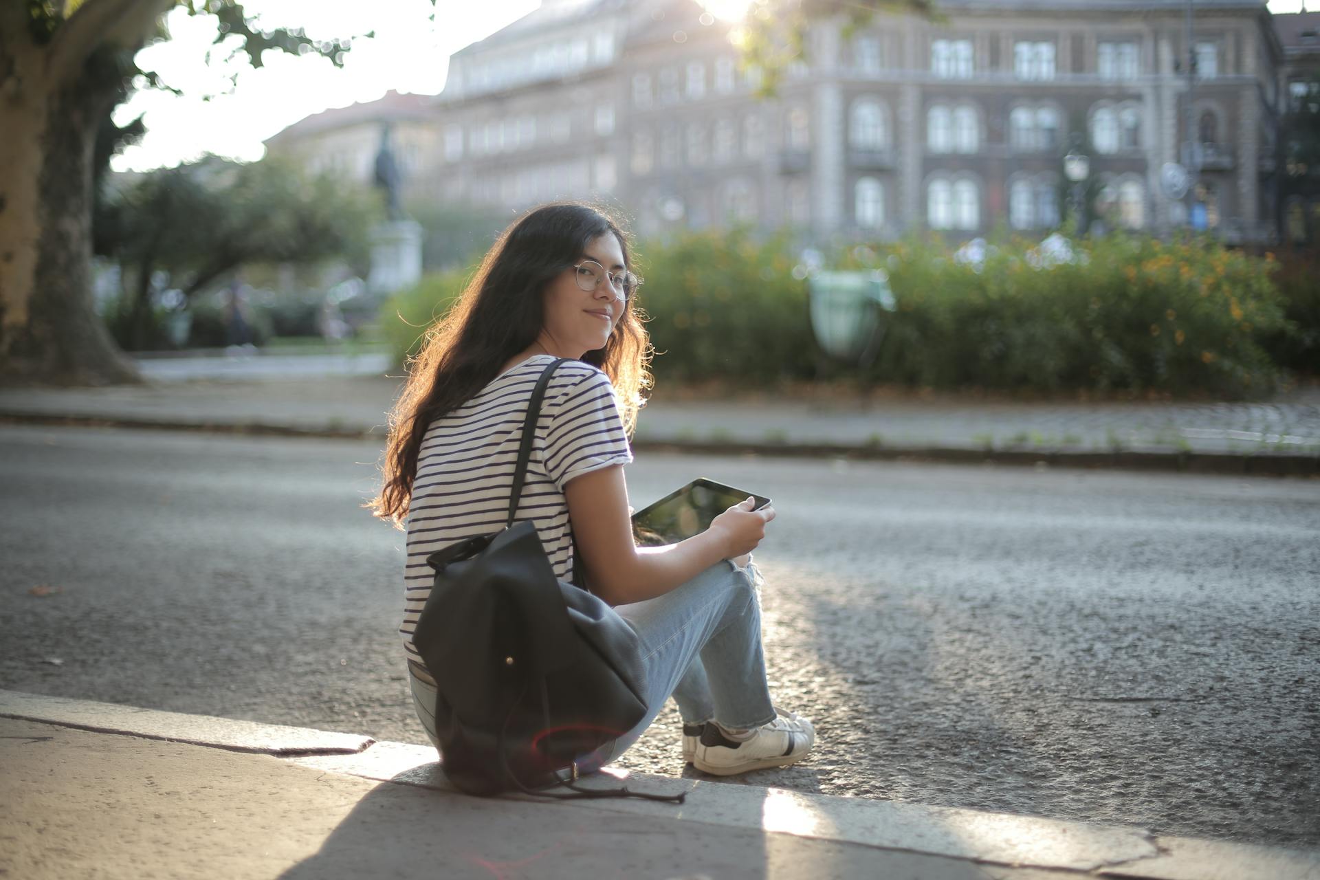 A smiling woman sitting outdoors on a sidewalk holding a tablet during the day.