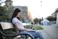 Woman in White and Black Stripe Shirt Sitting on Bench Using Laptop