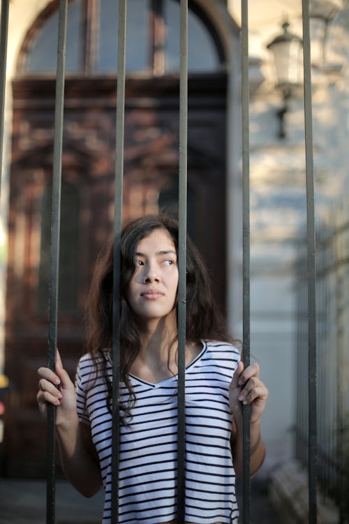 Free Curious isolated young woman looking away through metal bars of fence with hope at entrance of modern building Stock Photo