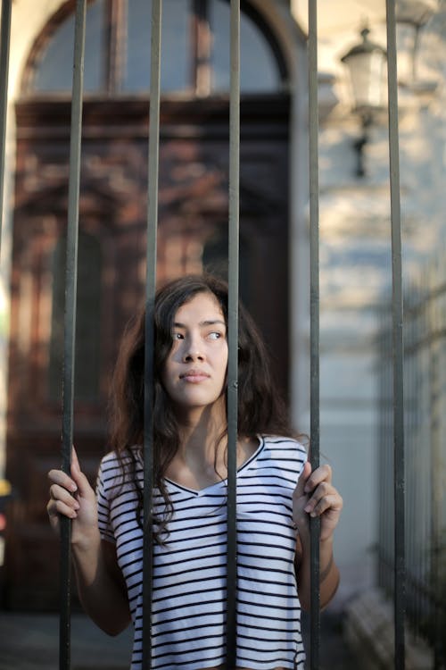 Curious isolated young woman looking away through metal bars of fence with hope at entrance of modern building