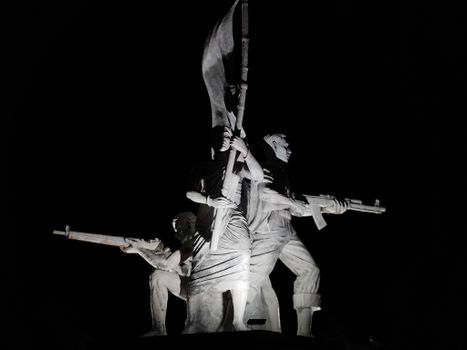 Striking night view of a monumental sculpture depicting armed figures holding a flag, symbolizing courage.