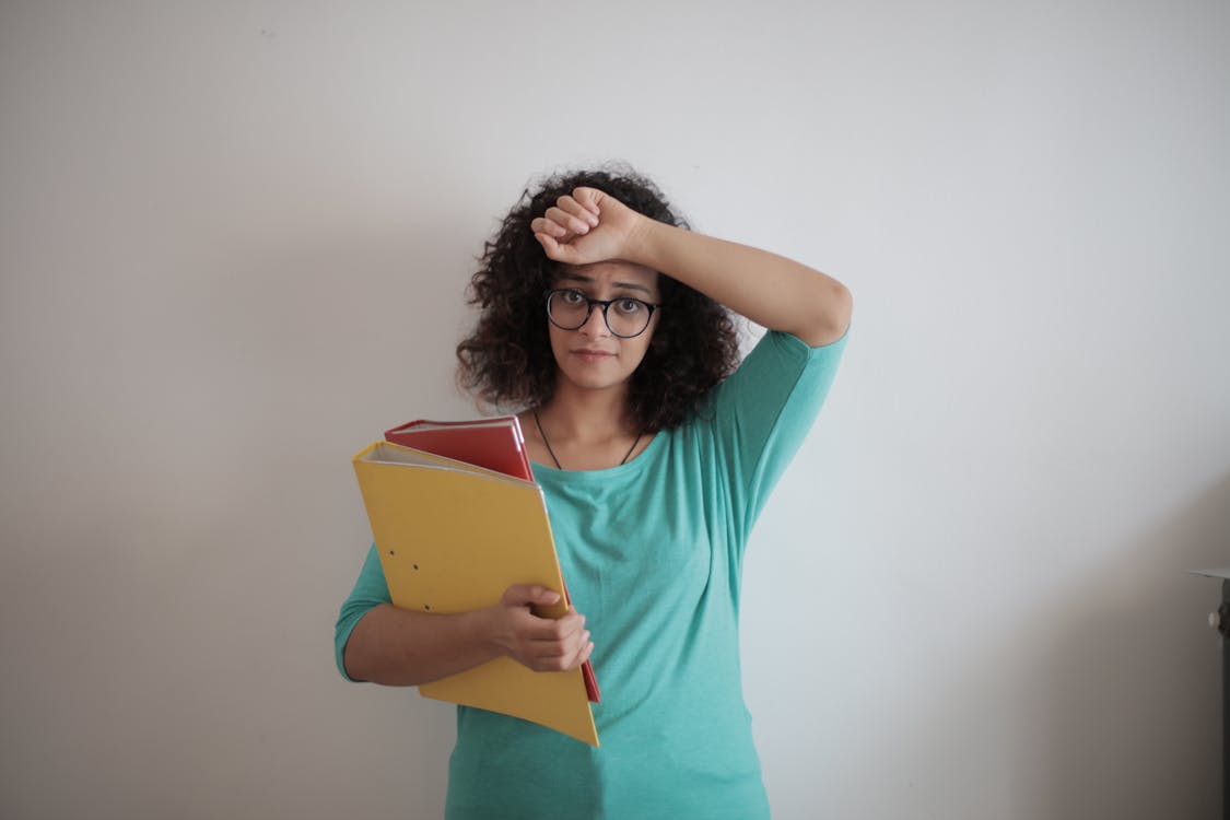 Unhappy overworked female employee in glasses and casual clothes with folders of documents looking at camera with sadness and touching head while standing against white wall in contemporary office