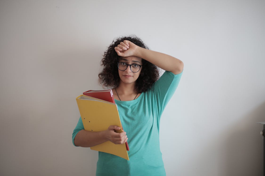 Free Unhappy overworked female employee in glasses and casual clothes with folders of documents looking at camera with sadness and touching head while standing against white wall in contemporary office Stock Photo