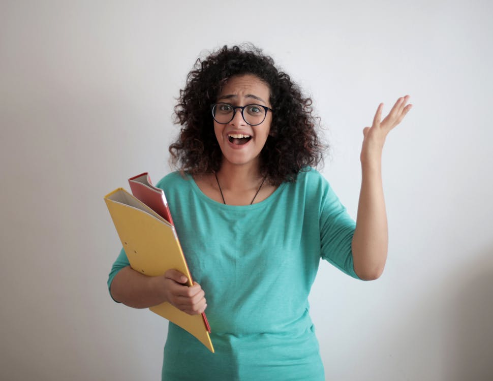 Free Dissatisfied annoyed woman with mouth opened wearing glasses and turquoise blouse looking away and screaming while standing against white wall with folders of documents and having problems in work Stock Photo