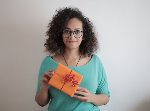Free Smiling woman with curly hair holding a vibrant orange gift with red ribbon on a neutral background. Stock Photo