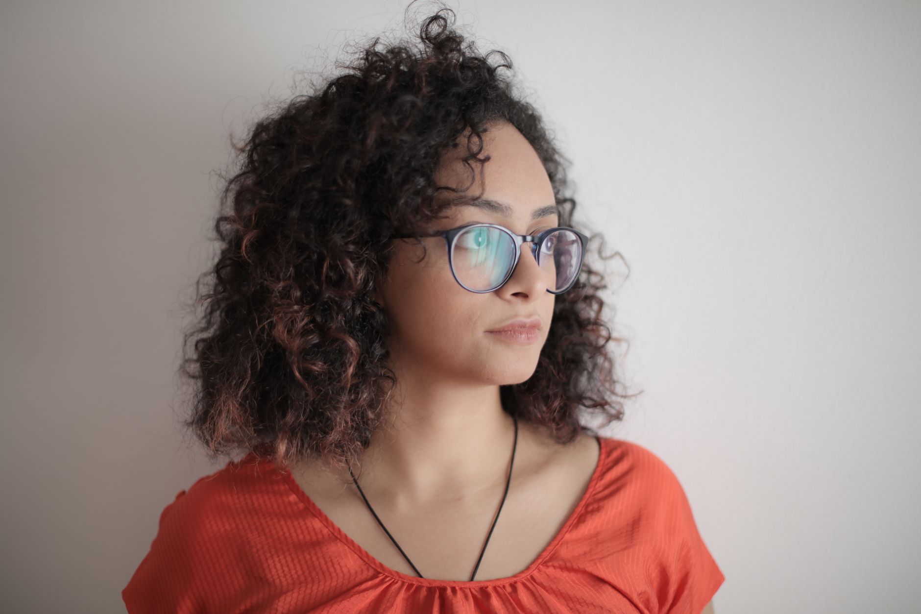 Thoughtful woman with curly hair and glasses in a red top against a white background.