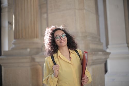 Free Low angle of gladful woman in round glasses and yellow shirt smiling at camera while standing with folder and backpack on street against blurred exterior of ancient building in old town Stock Photo