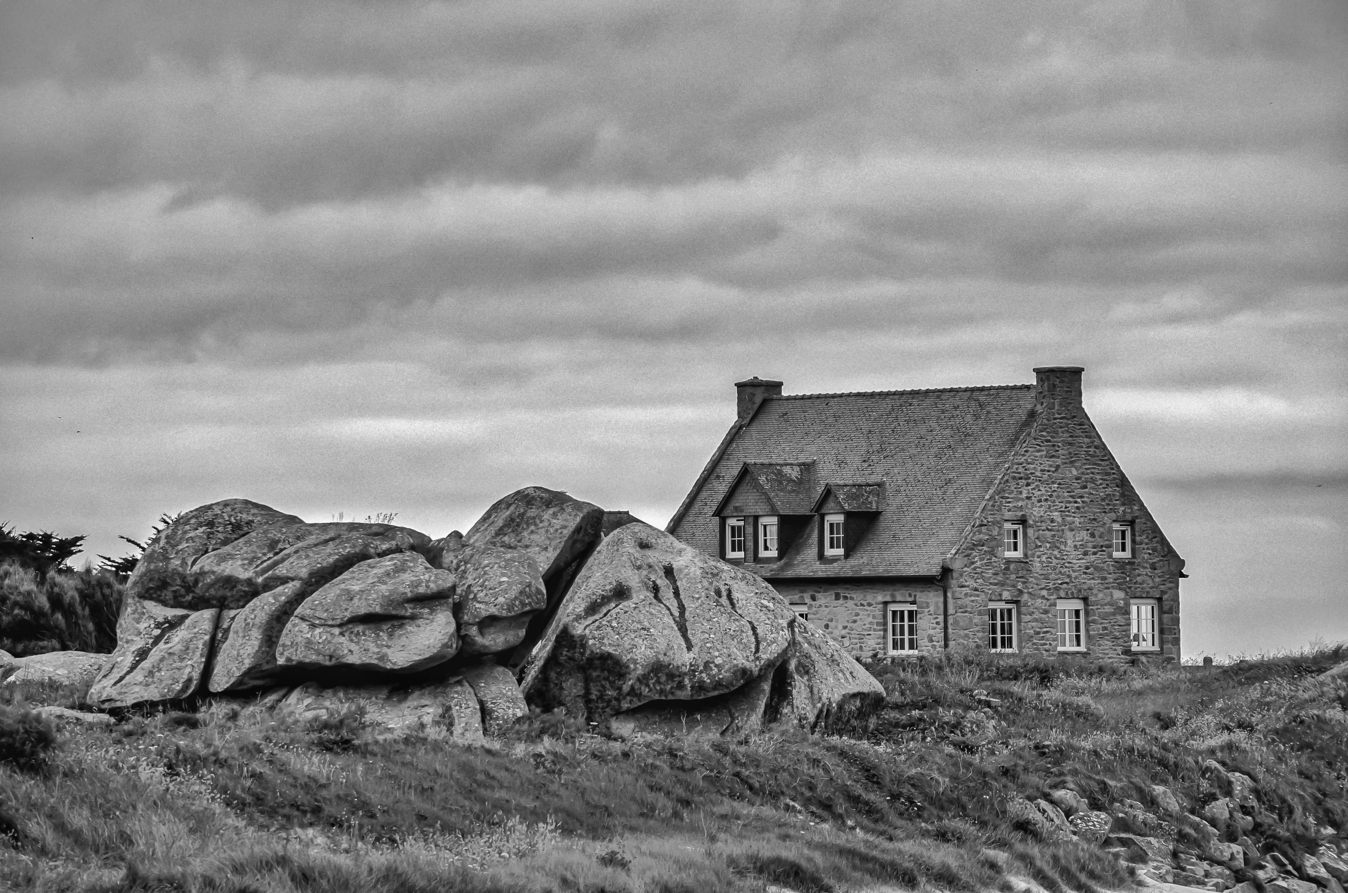A picturesque stone house sits near large boulders under a cloudy sky, evoking a sense of solitude and timeless beauty.