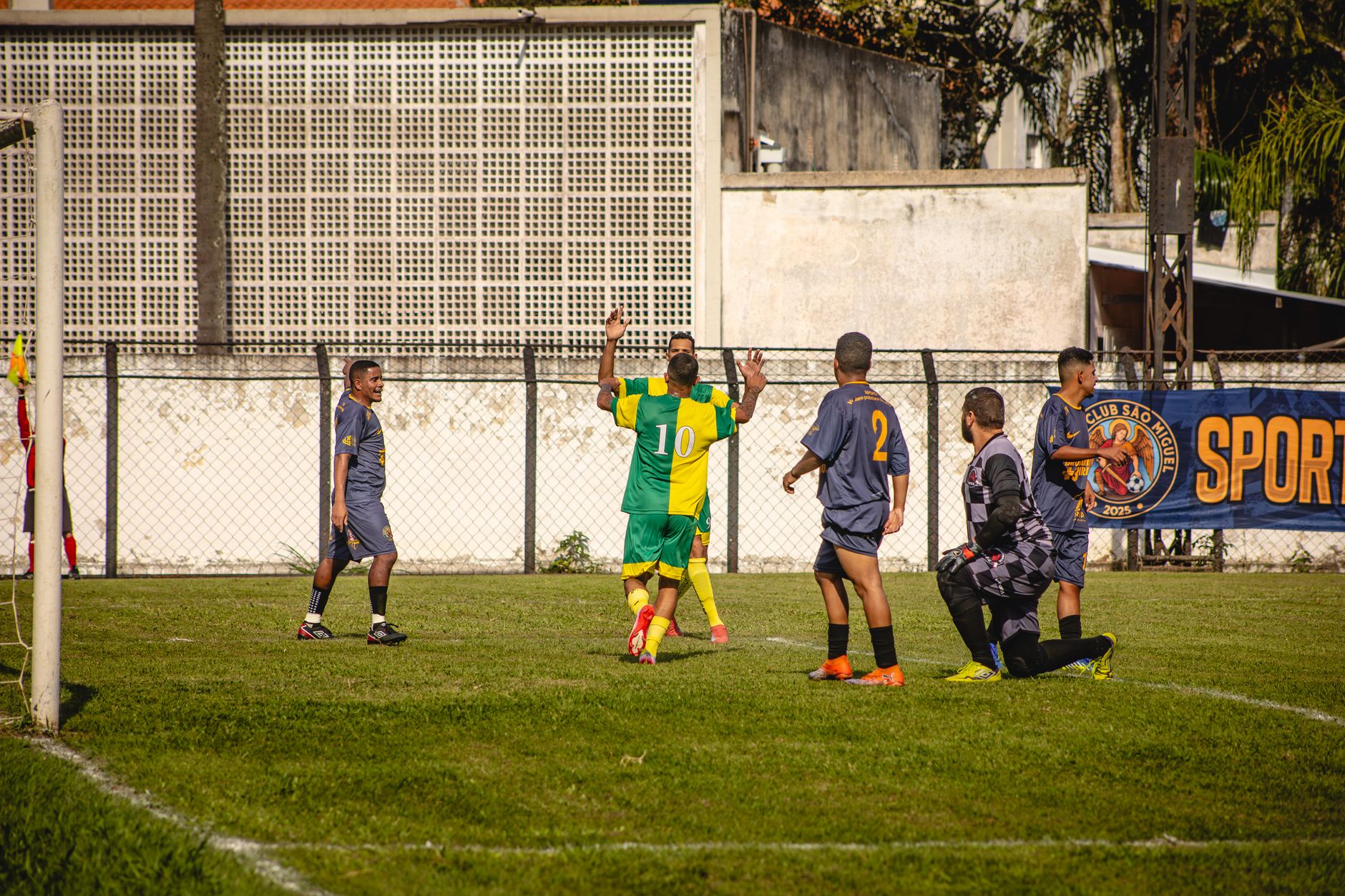 Amateur soccer match with players in green and yellow uniforms, outdoor setting on sunny day.
